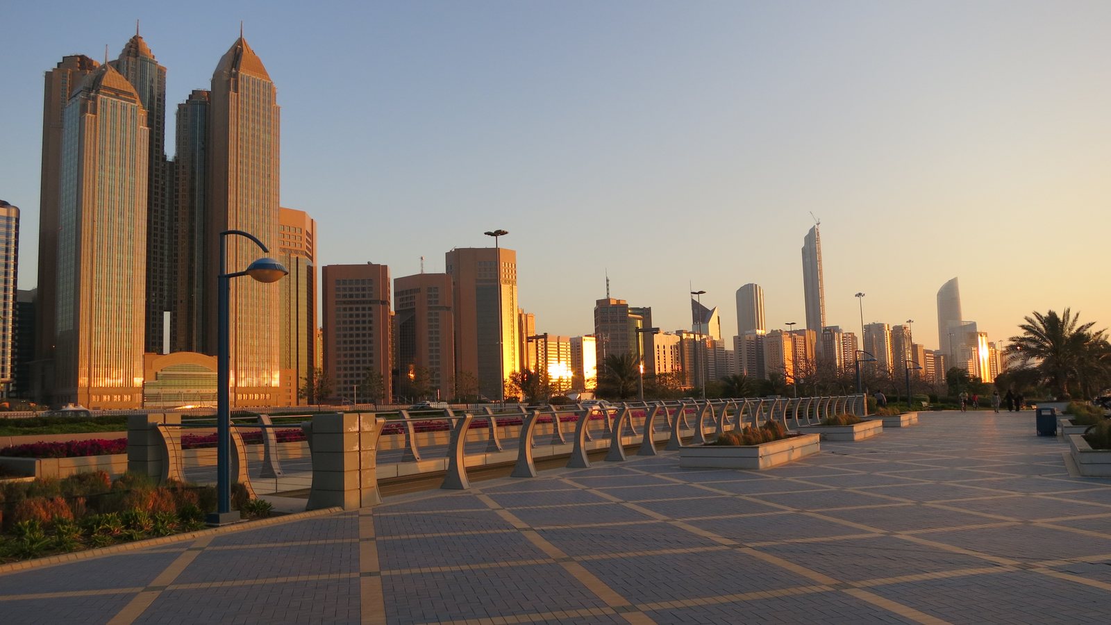 Abu Dhabi's Corniche skyline glowing gold at sunset, seen from street level on the promenade.