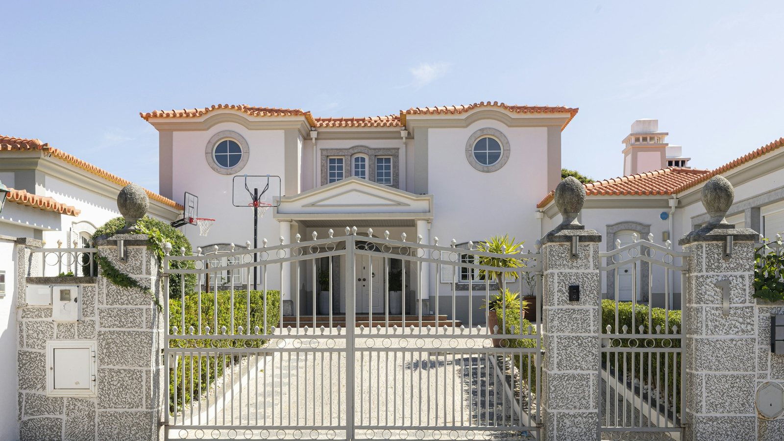 A large wrought-iron gate and stone gateposts closed across the driveway of a gated mansion, the house itself only partly visible behind.