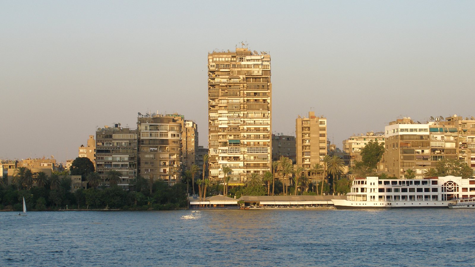 Cairo's Nile-side skyline glowing at sunset, seen across the river with a white Nile cruise boat moored at the corniche in the foreground.