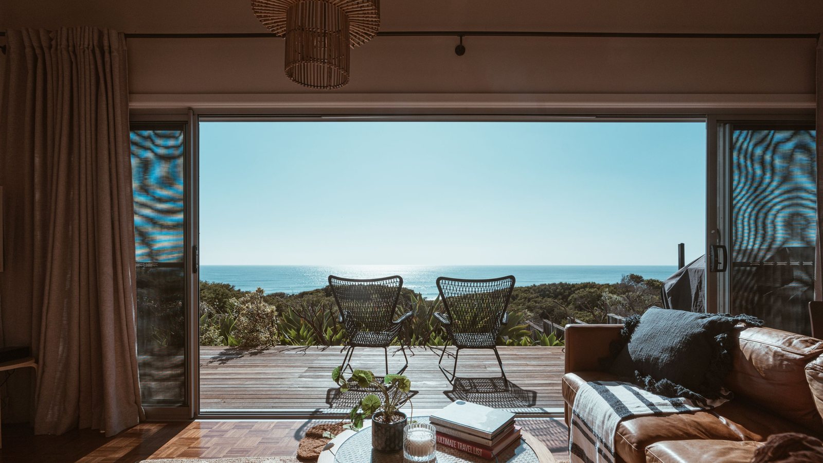 A modern villa living room opening through sliding doors onto a wood-decked terrace, looking straight out over the ocean.