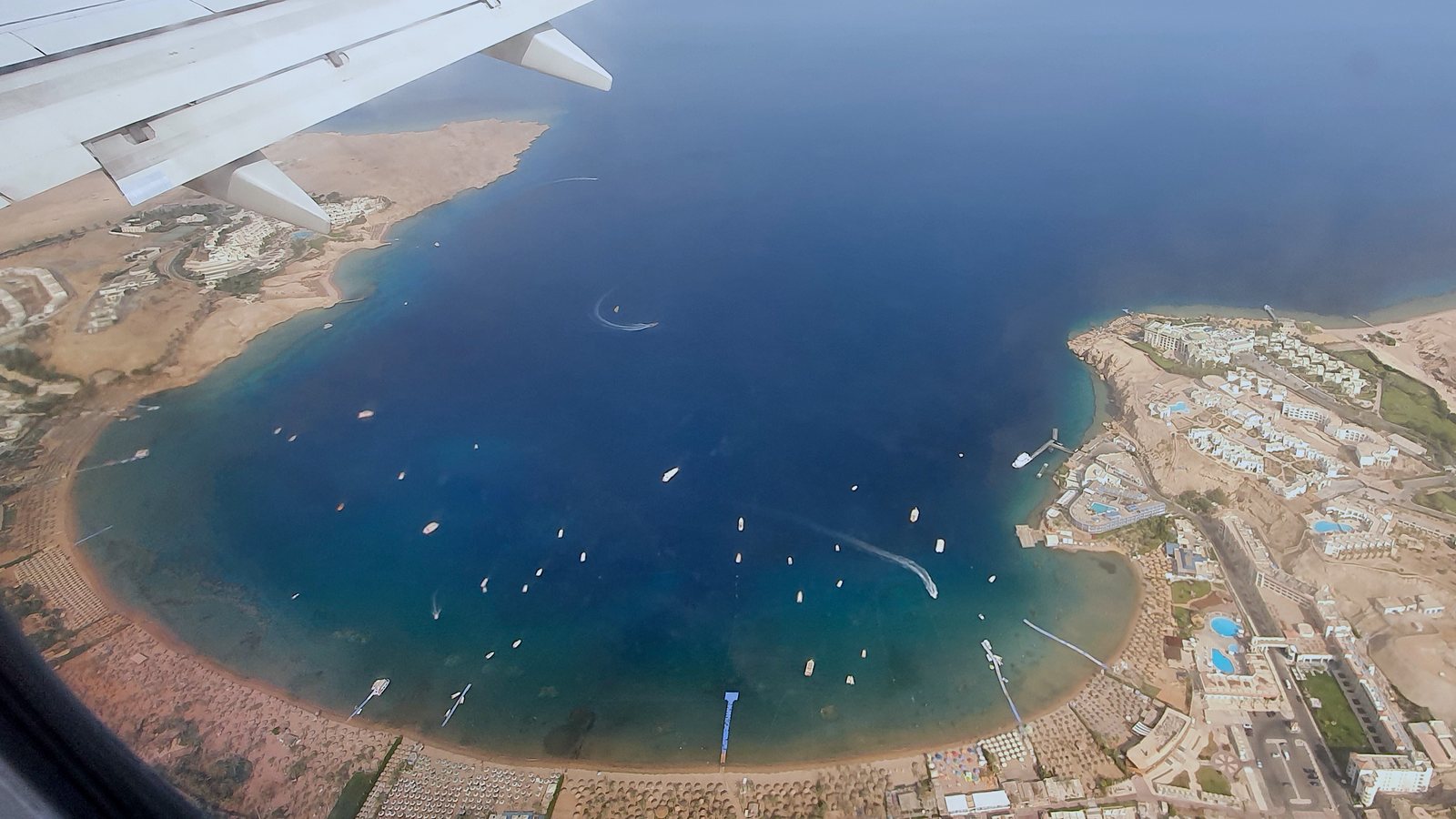 Aerial view of Naama Bay, Sharm El Sheikh, from a descending aircraft — turquoise Red Sea water ringed by resort hotels and beach umbrellas.